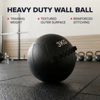 A black 3KG heavy duty medicine ball with a textured surface and reinforced stitching, resting on a gym floor with fitness equipment in the background. Text overlays highlight key features: 'HEAVY DUTY WALL BALL', 'TRAINING WEIGHT', 'TEXTURED OUTER SURFACE', and 'REINFORCED STITCHING'.